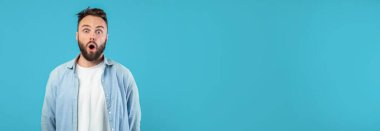Sakallı genç bir adam parlak mavi bir duvarın önünde duruyor, şaşkınlık ifadesi sergiliyor. O, günlük beyaz bir tişört ve düğmeli bir gömlek giyiyor, doğrudan izleyiciye bakıyor..