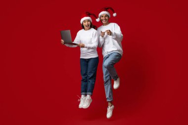 İki insan, tatil sezonunda heyecan ve neşe içinde Noel Baba şapkaları ve günlük giysiler giyip dizüstü bilgisayar tutarak parlak kırmızı bir arkaplana yaslanıyor..