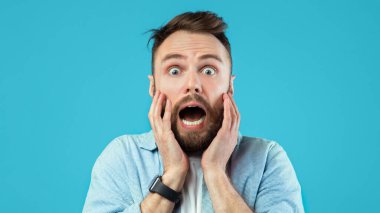 Sakallı bir adam iki eliyle yüzünü tutarken şaşırmış görünür. Gözleri tamamen açık ve beklenmedik bir şeye tepki veriyor gibi görünüyor. Arkaplan parlak mavi.