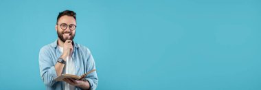 Elinde açık bir kitapla gülümseyen gözlüklü genç bir adam. Düşünceli görünüyor, rahat bir şekilde hafif bir gömlek giyiyor ve koyu mavi bir arka planın önünde duruyor, neşeli bir atmosfer yaratıyor..