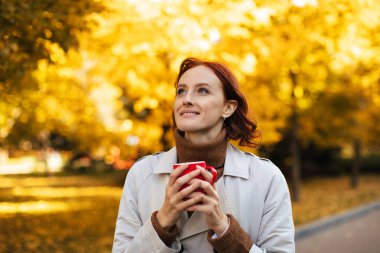 Altın yapraklı ağaçlarla çevrili manzaralı bir parkta duran bir kadın. Sıcak bir içki tutuyor ve gülümsüyor. Güz havasının ve güzel çevrenin tadını çıkarıyor..