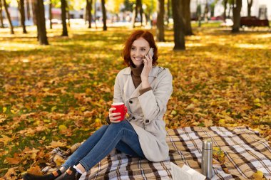 Renkli sonbahar yapraklarıyla dolu bir parkta bir kadın battaniyeye oturuyor. Elinde kırmızı bardakla telefonuyla konuşuyor. Güneşli bir günde mutlu ve rahat görünüyor..