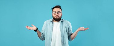 Gözlüklü ve sakallı bir adam sağlam bir mavi zemine karşı duruyor, kafası karışmış bir ifadeyle omuzlarını silkiyor. Açık renk tişört ve tişört giyiyor..