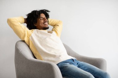 Genç siyah bir adam evinde rahat bir koltukta dinleniyor, gülümsüyor ve boş zamanlarının tadını çıkarıyor. Elleri başının arkasında oturur, huzurlu bir ortamda neşe ve neşe saçar..