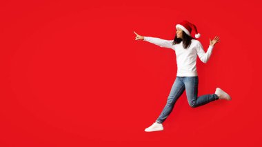 Noel Baba şapkası takan neşeli bir kadın canlı kırmızı bir arka plana işaret ederken şakacı bir şekilde sıçrıyor. Sahne, tatil sezonunun heyecanını yakalıyor..