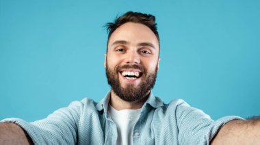 Sakallı bir adam selfie çekerken geniş ölçüde gülümser. Açık renk bir gömlek giyiyor ve parlak mavi bir duvarın önünde duruyor. Neşeli bir atmosfer yaratıyor..