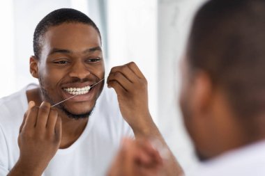 Genç bir adam parlak bir banyoda dişlerini temizlemek için diş ipi kullanırken gülümsüyor. Sahne, sabah rutini sırasında bir anlık kişisel bakım görüntüsü yakalıyor..