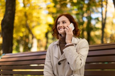 Genç bir kadın parkta bir bankta oturuyor, telefon konuşmasından zevk alıyor. Arka planda canlı sonbahar yaprakları var, sıcak ve davetkar bir atmosfer yaratıyor..