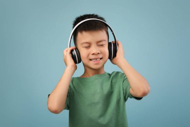 Büyük kulaklıklardan müzik dinlerken gözleri kapalı bir çocuk gülümsüyor. Sakin bir ortamda yeşil bir gömlek giyerken neşeli ve rahat görünüyor..