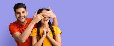 Genç bir adam, gülümseyen genç bir kadının gözlerini şakacı bir şekilde kapatarak eğlenceli bir an yaratıyor. Parlak mor bir arka planda dururken heyecan ve merak gösteriyor..