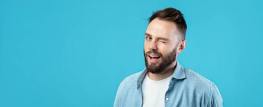 Sakallı bir adam gülümsüyor ve beyaz bir tişörtün üzerine açık mavi bir gömlek giyiyor. Parlak mavi arka plan, sahneye canlı bir dokunuş ekler..