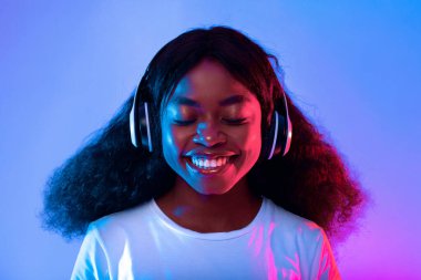 Kıvırcık siyah saçlı bir kadın kulaklık takıyor, gözleri kapalı gülümsüyor. Canlı mavi ve mor ışıklar neşeli bir atmosfer yaratarak onun neşeli yüz ifadesini güçlendirir..