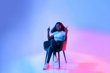 Genç bir kadın heyecan içinde modern bir sandalyede otururken gülümsüyor. Parlak mavi ve pembe ışıklar etrafında canlı bir atmosfer yaratır..