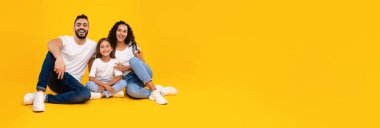 Neşeli bir aile, canlı sarı bir arka planda birlikte oturur. Ebeveynler ve küçük kızları, güçlü bağlarını ve mutluluklarını göstererek gülümsüyorlar..