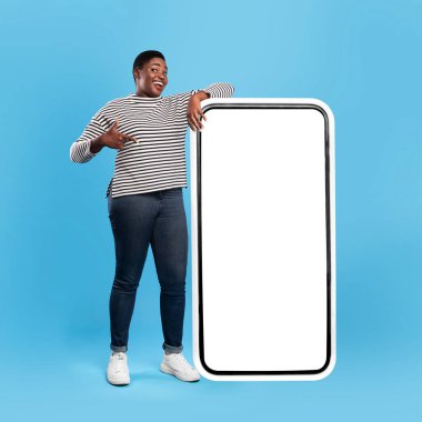 Çizgili tişörtlü genç bir adam büyük boş bir ekranın yanında duruyor, parlak mavi bir stüdyoda heyecandan gülümsüyor ve el sallıyor. Çevre hareketli ve eğlenceli..