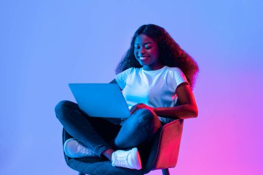 Genç bir kadın bir sandalyede rahatça otururken dizüstü bilgisayarını kullanırken gülümsüyor. Sahne canlı bir atmosfer yaratan parlak mavi ve pembe ışıkla güçlendirilir..