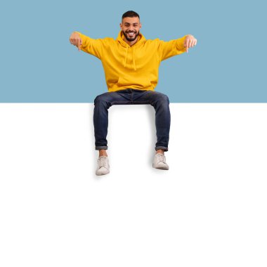 Sarı kapüşonlu bir adam kenarda oturuyor, neşeli ve enerjik görünüyor. Arka plan yumuşak mavi bir renge sahip, canlı ve neşeli bir atmosfer yaratıyor..