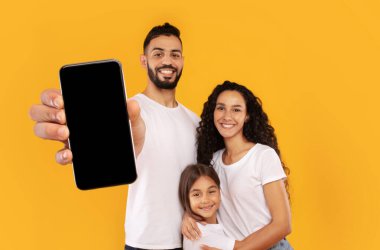 Üç kişilik bir aile, canlı sarı bir arkaplanın önünde duruyor ve gülümsüyor. Baba akıllı bir telefon tutarken, anne ve kız kucaklaşıyor, sıcaklığını ve mutluluğunu gösteriyor..