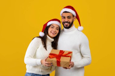 Paketlenmiş bir hediyeyle gülümseyen genç bir çift. Kış süveteri ve Noel Baba şapkalarını parlak sarı bir arka plana takıp neşeli bir tatil ruhunu yakalıyorlar..