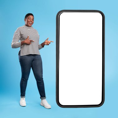 Neşeli bir kadın, parlak mavi bir ortamda büyük, boş bir telefon ekranının yanında duruyor. Çizgili bir kazak, kot pantolon ve spor ayakkabı giyiyor. İki eliyle neşeli bir poz veriyor..
