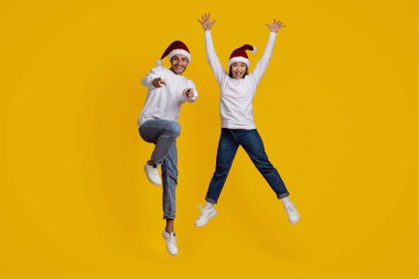 İki neşeli arkadaş havaya atlarken Noel şapkası ve beyaz kazak giyerler. Parlak sarı bir arka plana karşı mutluluğu ifade ederler, birlikte tatil ruhunu kutlarlar..