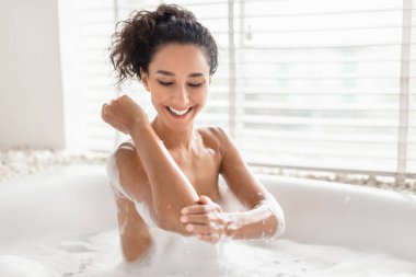 Bir kadın köpük banyosu yaparken kolunu sabunla ovalarken gülümsüyor. Güneş ışığı pencereden süzülür ve banyoda sıcak ve sakin bir atmosfer yaratır..