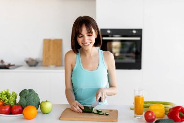 Parlak bir mutfakta, kolsuz tişörtlü bir kadın kesim tahtasındaki salatalıkları seve seve doğruyor. Renkli meyve ve sebzeler onu çevrelemiş, bir bardak meyve suyu ile birlikte, sağlıklı bir yaşam tarzını yansıtıyor..