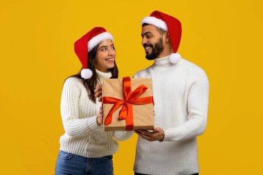 Kırmızı Noel Baba şapkası takan neşeli bir çift kırmızı kurdeleyle bağlanmış güzel bir hediyeyi tutarken gülümsüyor. Onların rahat beyaz kazakları neşeli ruhu tamamlıyor..