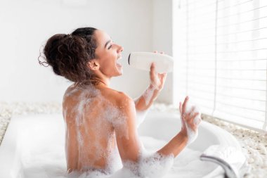 Bir kadın köpükle dolu köpük banyosunda gevşiyor, elinde bir şişe tutuyor ve neşeyle gülüyor. Güneş ışığı pencereden akar ve dingin atmosferi güçlendirir..