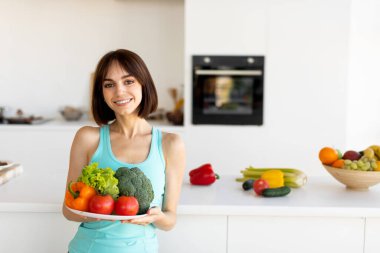 Parlak mutfak sahnesinde elinde taze sebze ve meyve tabağı tutarken gülümseyen mavi kolsuz bir kadın vardı. Doğal ışık odayı dolduruyor, sağlıklı seçimleri vurguluyor..