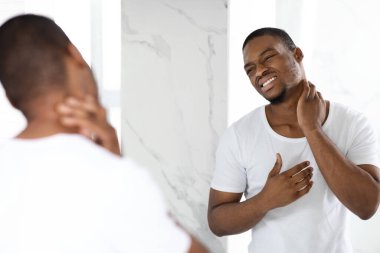 Beyaz tişörtlü bir adam aynanın önünde duruyor, parlak bir banyoda sabah rutinini yaparken boyun ağrısı belirtileri gösteriyor. Onun ifadesi rahatsızlık ve endişeyi yansıtıyor..