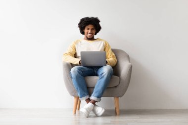 Yakışıklı, genç Black serbest çalışan evdeki rahat koltuğunda oturup dizüstü bilgisayarıyla çalışmaya odaklanmış. Etrafı sakin bir atmosferle çevriliyken uzak işlerin esnekliğini seviyor..