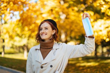 Genç bir kadın sonbaharda bir parkta yüz maskesi tutarken gülümser. Canlı sarı ve turuncu yapraklar neşeli bir ortam yaratmak için sıcak bir zemin oluşturur..