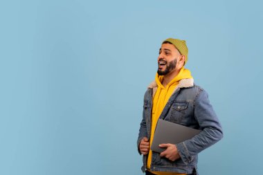 Genç bir adam seve seve dizüstü bilgisayar tutuyor, sarı kapüşonlu ve kot ceketli. Açık mavi bir arkaplanda duruyor, neşeli ve rahat bir atmosferi taşıyor..