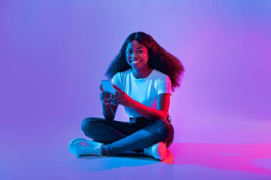 Genç bir kadın yerde bağdaş kurmuş oturuyor, akıllı telefonunu kullanırken gülümsüyor. Canlı mor ve mavi ışık etrafında canlı bir atmosfer yaratıyor..