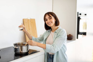 Parlak ve modern bir mutfakta, bir kadın ocakta bir tencere karıştırır, yemek pişirirken mutlu bir şekilde gülümser. Mutfak ahşap kaplar ve mutfak malzemeleriyle güzelce düzenlenmiş..