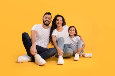 Neşeli bir aile, parlak sarı bir arka planda yerde yan yana oturur. Eşleşen beyaz gömlekler ve kotlar giyip neşeli gülüşler atıyorlar..