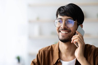 Gözlüklü genç bir adam, aydınlık bir odada akıllı telefonuyla konuşurken gülümsüyor. Arkaplanda açık renkli raflar ve yeşillikler var. Neşeli bir atmosfer yaratıyorlar..