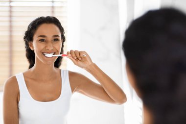 Dişlerini fırçalarken gülümseyen bir kadın banyo aynasının önünde duruyor. Parlak uzayın pencereden gelen doğal bir ışığı var. Bu an, onun sabah rutinini yakalıyor..