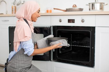 Açık pembe tesettürlü bir kadın, parlak ve modern bir mutfakta dururken fırına dikkatlice bir yemek kabı koyar. Mutfakta temiz çizgiler ve nötr renkler var..