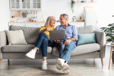 İki son sınıf öğrencisi kanepede rahatça oturur, gülümser ve dizüstü bilgisayara bakarlar. Günışığı modern oturma odasını dolduruyor. Sıcak ve davetkar bir atmosfer yaratıyor..