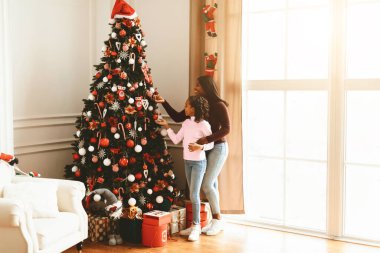 İki aile üyesi oturma odalarında bir Noel ağacı süsler. Ağaç renkli süslemelerle kaplı ve hediyelerle çevrili. Güneş ışığı pencereden girer..