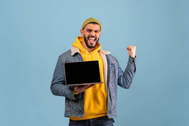 Kocaman gülümseyen genç bir adam dizüstü bilgisayarını tutarken yumruğunu kaldırıyor. Açık mavi arka planda sarı kapüşonlu ve kot ceketli sıradan bir kıyafet giyiyor..
