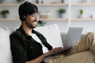 Genç bir adam bir kanepede rahatça otururken dizüstü bilgisayarında çalışırken gülümsüyor. Rahat bitkiler ve şık bir iç mekan rahat bir ev ortamını yansıtır..