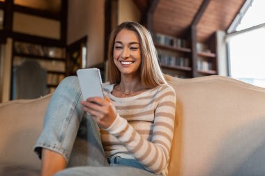 Genç bir kadın açık renkli bir kanepede rahat bir şekilde oturuyor, akıllı telefonuyla meşgul olurken gülümsüyor. Samimi odada kitap dolu ahşap kirişler ve raflar bulunuyor..