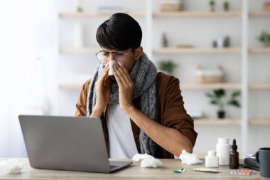 Genç bir adam ofisteki bir masada oturuyor, burnunu mendille silerken iyi görünmüyor. Etrafına çeşitli soğuk algınlığı ilaçları saçılıyor..