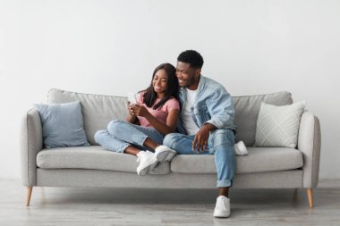 Birlikte vakit geçirmek. Akıllı telefonuyla sosyal medyayı paylaşan mutlu zenci kadın portresi gülümseyen erkek arkadaşıyla koltukta oturan çift, evdeki oturma odasında dinlenen iki arkadaş.