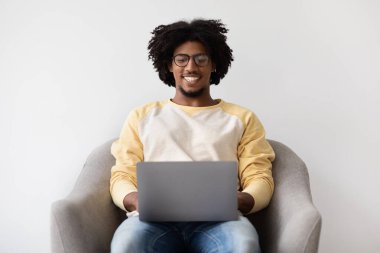 Genç siyah gözlüklü bir erkek evdeki koltukta rahatça oturur ve dizüstü bilgisayarıyla meşgul olur. Uzaktan çalışırken mutlu ve odaklanmış görünüyor..