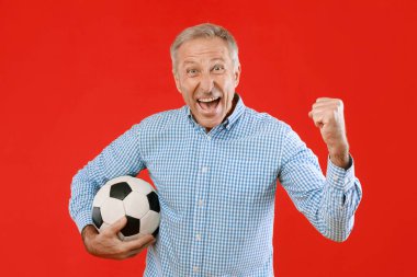 Bir adam bir futbol topu tutuyor ve büyük bir gülümsemeyle yumruğunu kaldırarak heyecan gösteriyor. Arka plan kırmızı, sahnenin coşkusunu arttırıyor..
