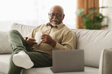 Son sınıf öğrencisi rahat bir oturma odasında kanepede oturur, kahve içerken gülümser ve dizüstü bilgisayarına bakar. Doğal ışık boşluğu doldurur ve sıcak bir atmosfer yaratır..